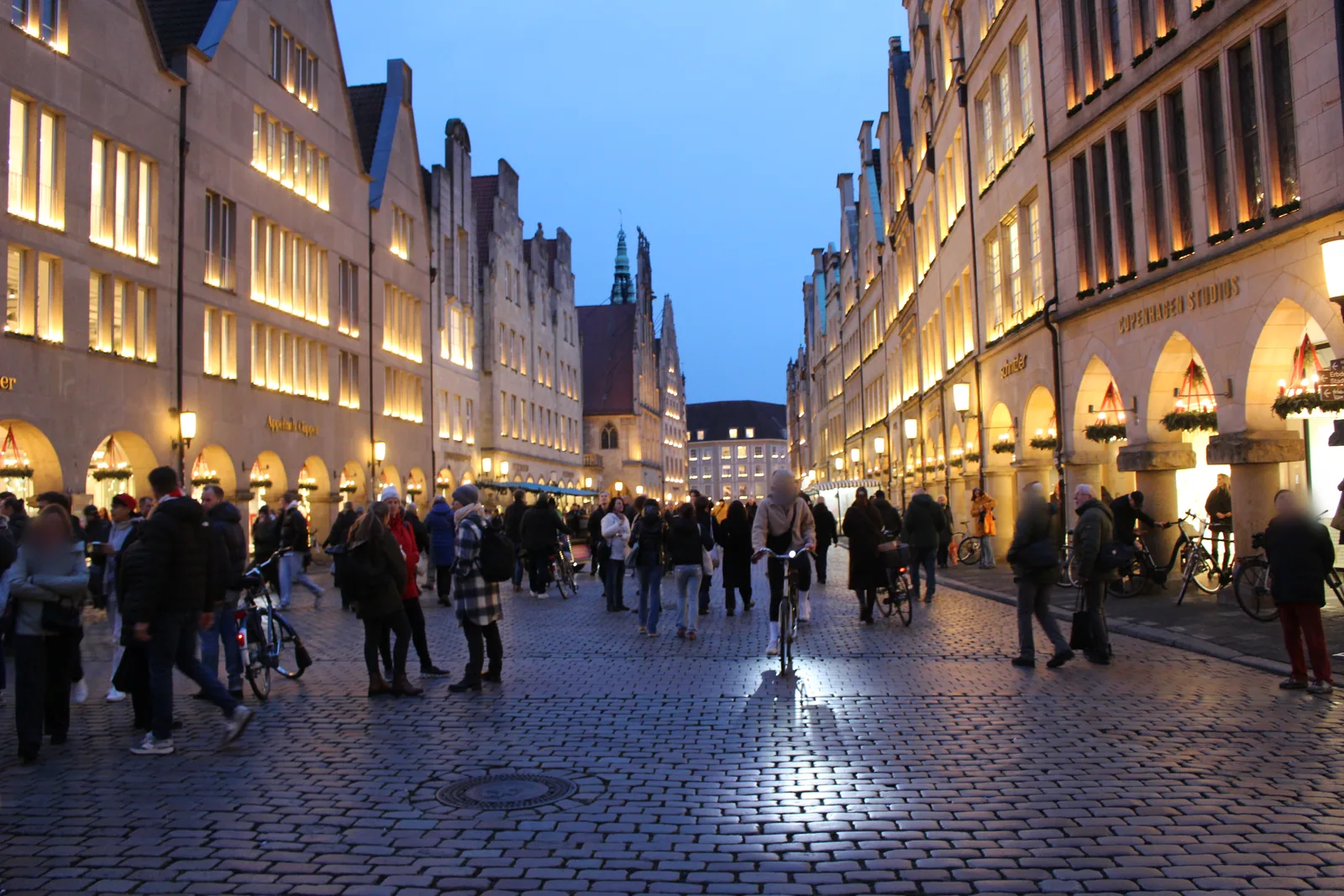 Prinzipalmarkt in Münster zur blauen Stunde: warm beleuchtete Giebelhäuser und Bogengänge mit Adventskränzen, Spaziergänger und Radfahrer auf dem regennassen Kopfsteinpflaster.