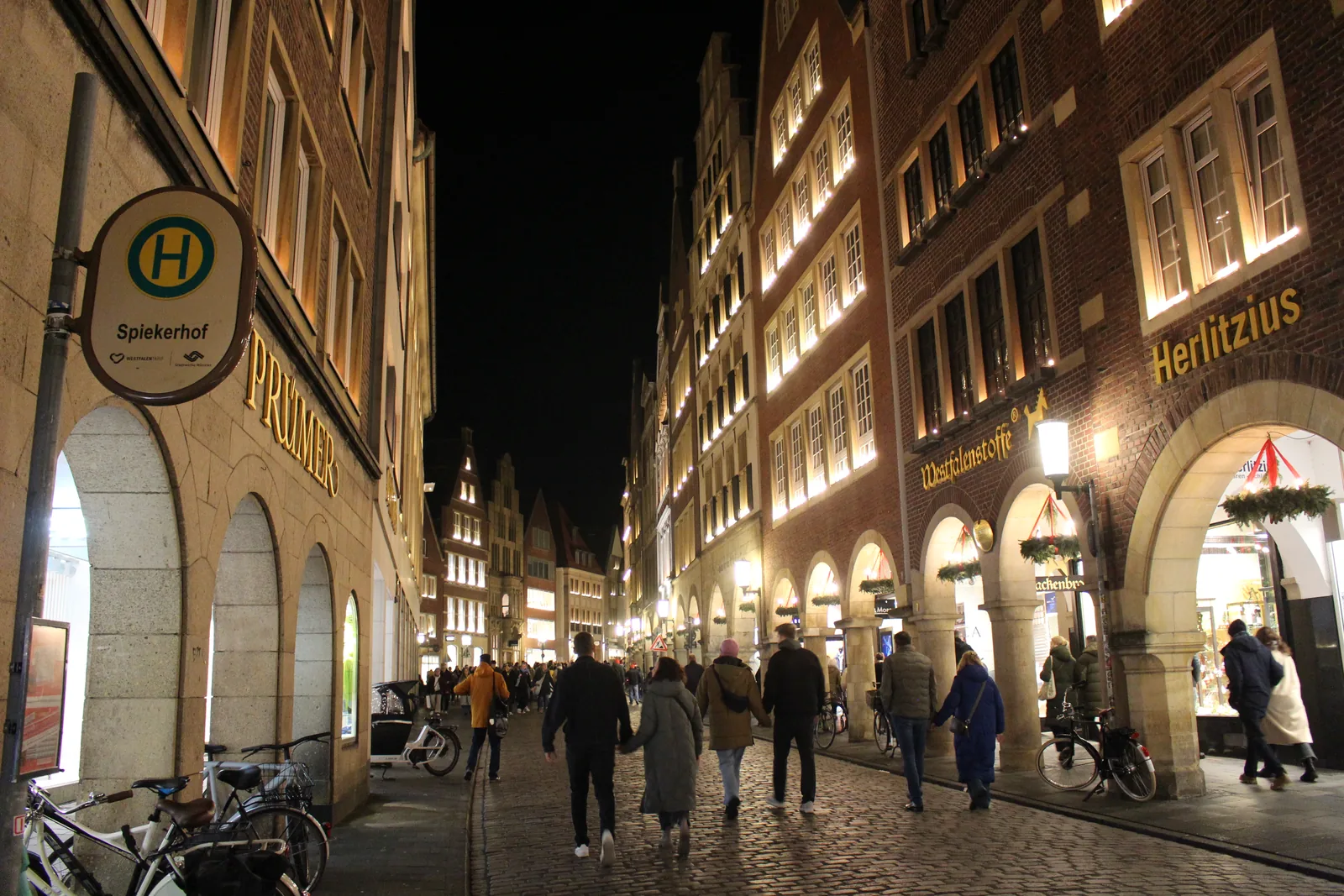Abendlich beleuchtete Giebelhäuser und Bogengänge mit Adventskränzen am Roggenmarkt in Münster, Spaziergänger auf dem Kopfsteinpflaster, vorne das Haltestellenschild Spiekerhof.