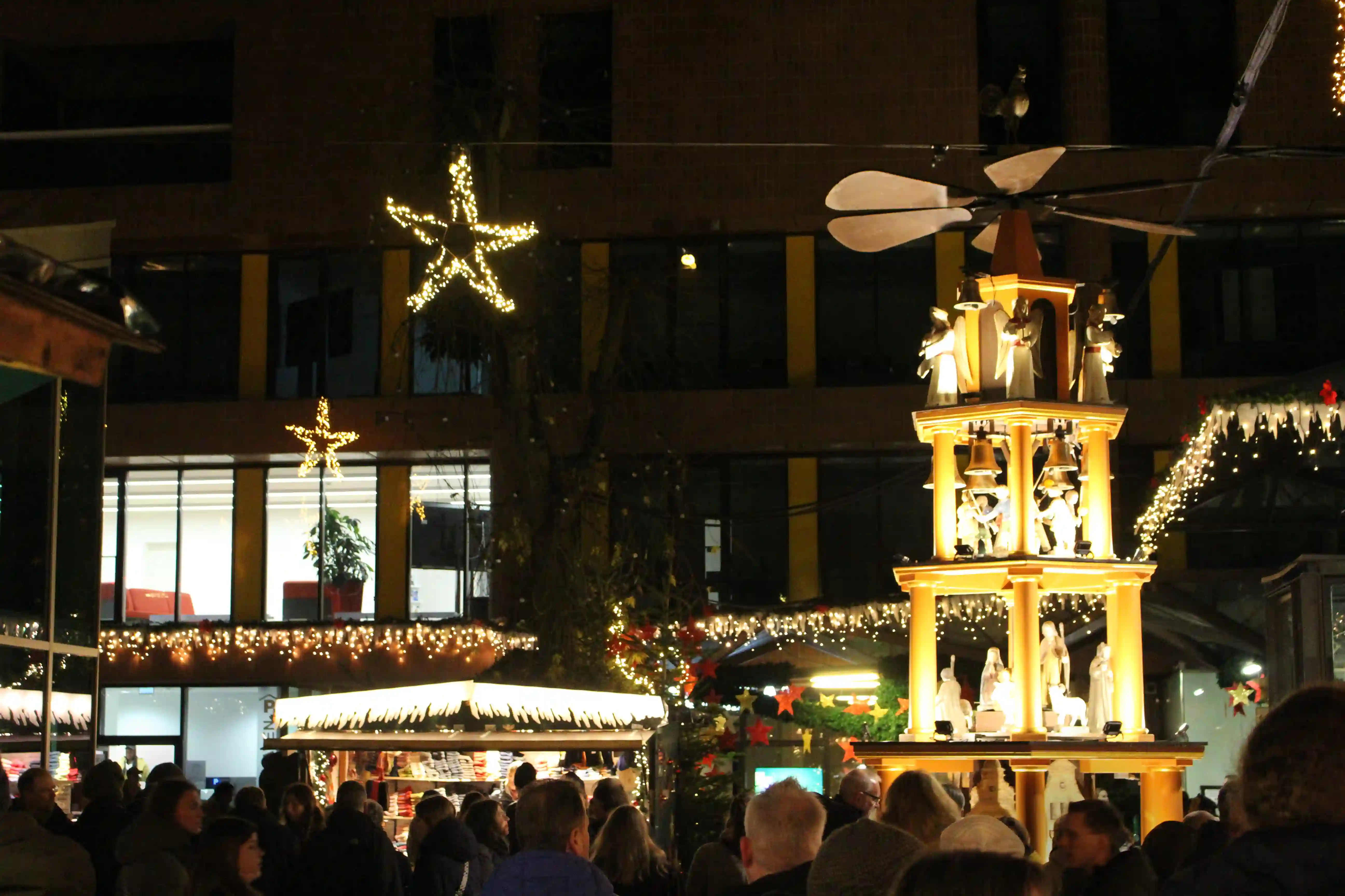 Große beleuchtete Holzpyramide mit Flügelrad, Engeln und Glocken auf dem Aegidii-Weihnachtsmarkt in Münster, über der Besuchermenge im Innenhof.