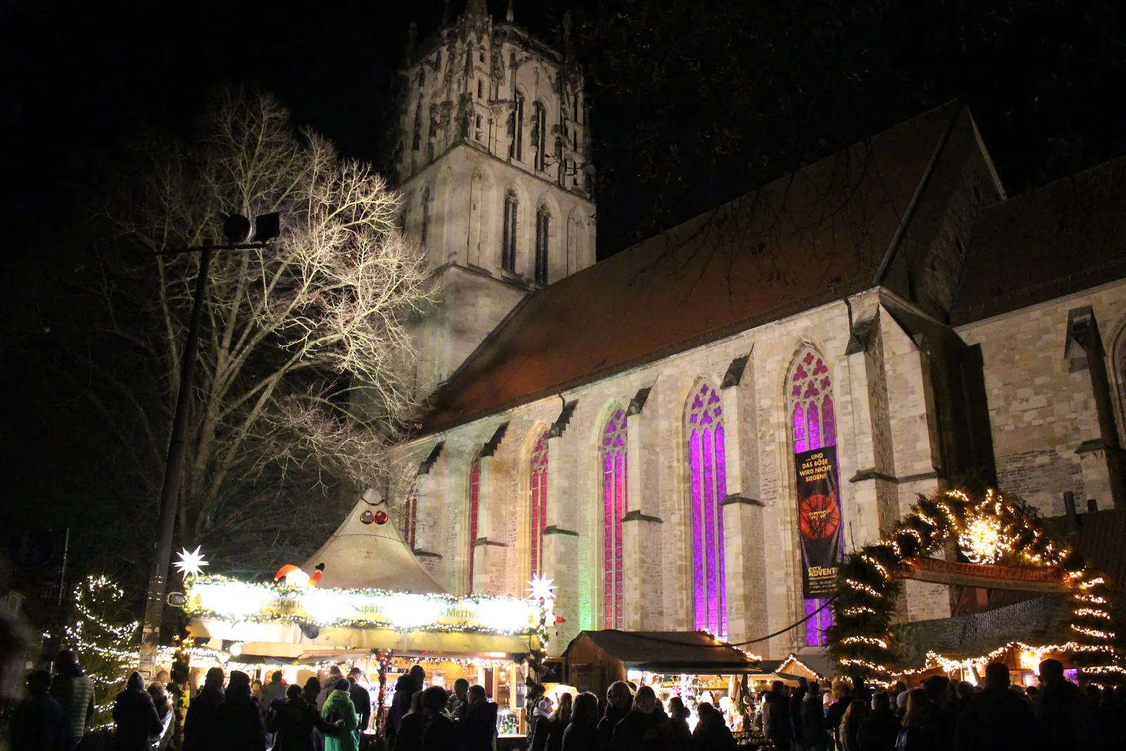 Giebelhüüskesmarkt vor der abends bunt beleuchteten Liebfrauen-Überwasserkirche in Münster, mit Buden und Menschen.