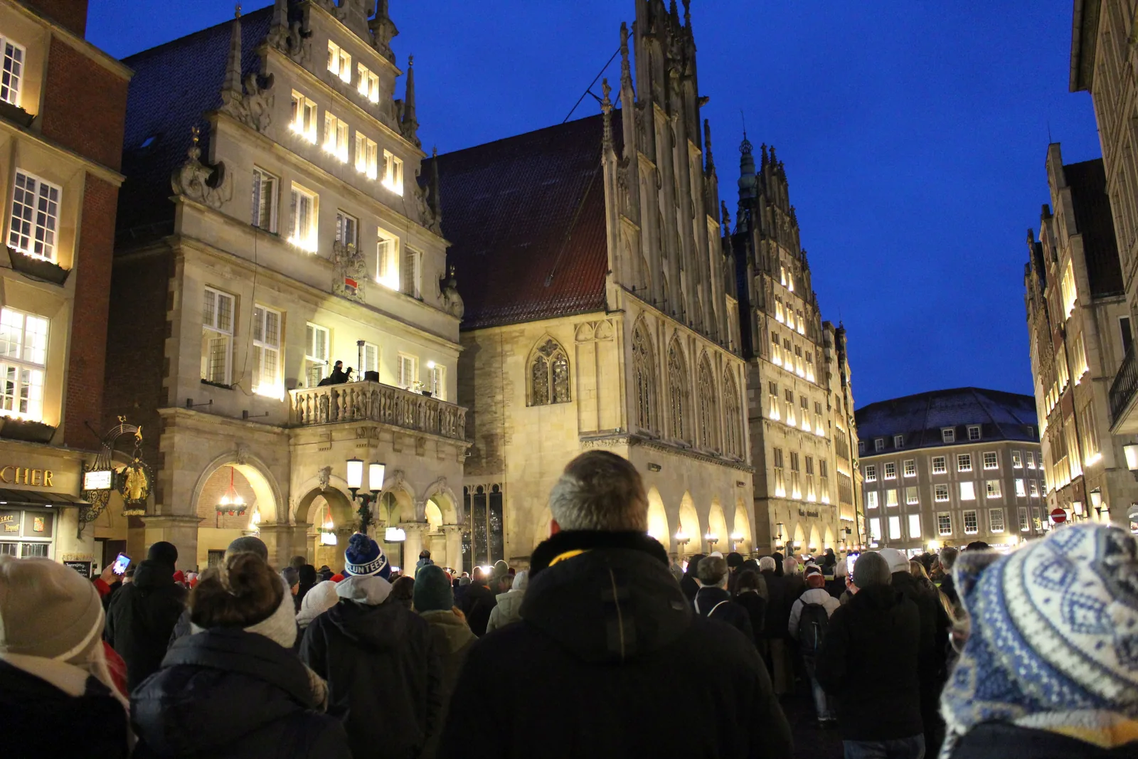 Adventsmusik vom Balkon des Stadtweinhauses am Prinzipalmarkt in Münster: Zur blauen Stunde hören viele Menschen zu, daneben das beleuchtete Historische Rathaus mit seinem gotischen Giebel.